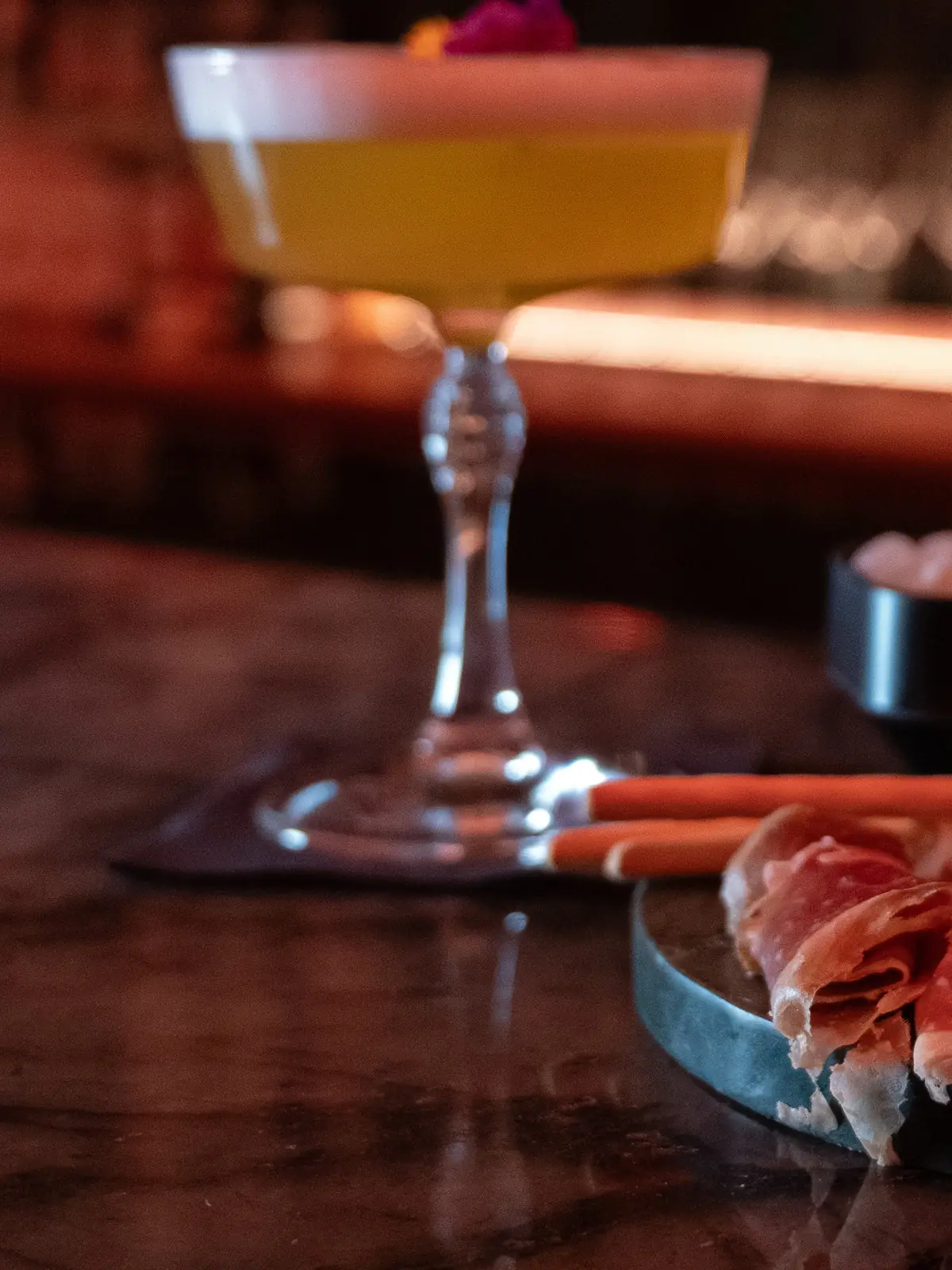 A cocktail in a coupe next to a plate of ham on the bar at Mayfair